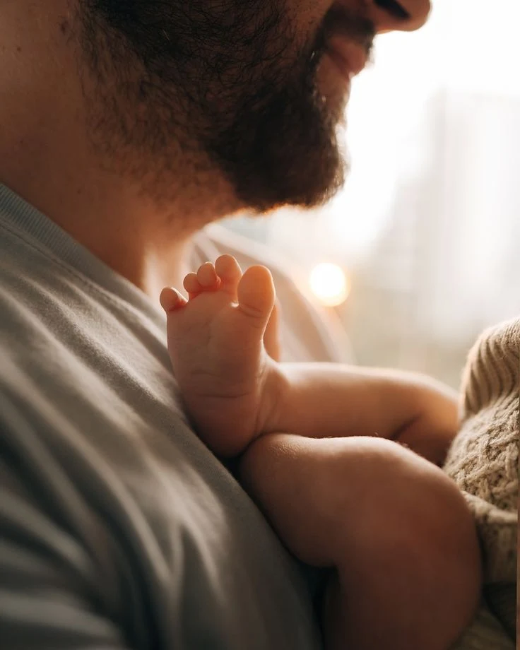 A father's hands gently holding his baby's tiny bare feet on cream muslin.